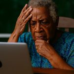 An elderly African American woman sits at a wooden table during the day, staring at a silver laptop with a puzzled and slightly frustrated expression. She wears a vibrant blue and green patterned blouse and gold earrings, with short silver curls framing her face. One hand rests on her cheek, the other touches her forehead. The soft natural light highlights her furrowed brow and thoughtful gaze. Behind her, a window reveals lush greenery, while the desk around her is cluttered with everyday items. The image captures the emotional tension of navigating unfamiliar technology with dignity and grace