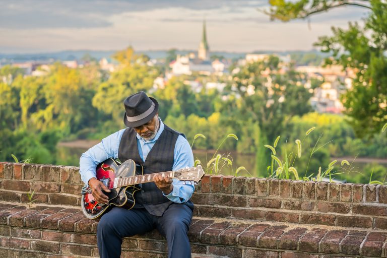 A Black elder sitting on a brick wall playing a guitar. Finally living life his way.