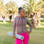 An African American senior lady with a yoga mat with palm trees in the background.