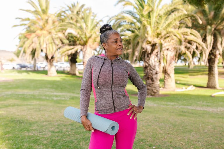 An African American senior lady with a yoga mat with palm trees in the background.